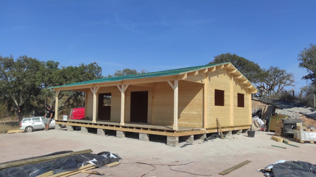 Casa de madeira maciça com alpendre corrido em construção, entre sobreiros, com membrana verde na cobertura