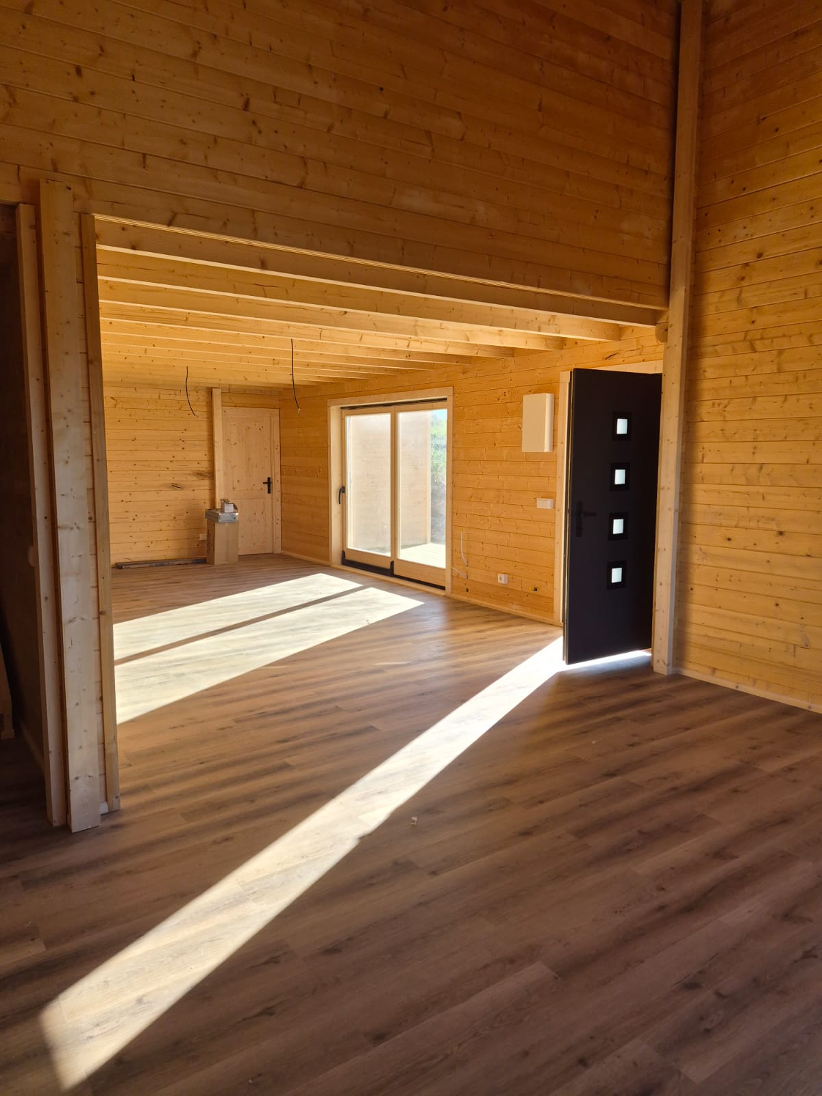 Sala da casa de madeira com pavimento laminado, porta de entrada e porta de correr para o alpendre