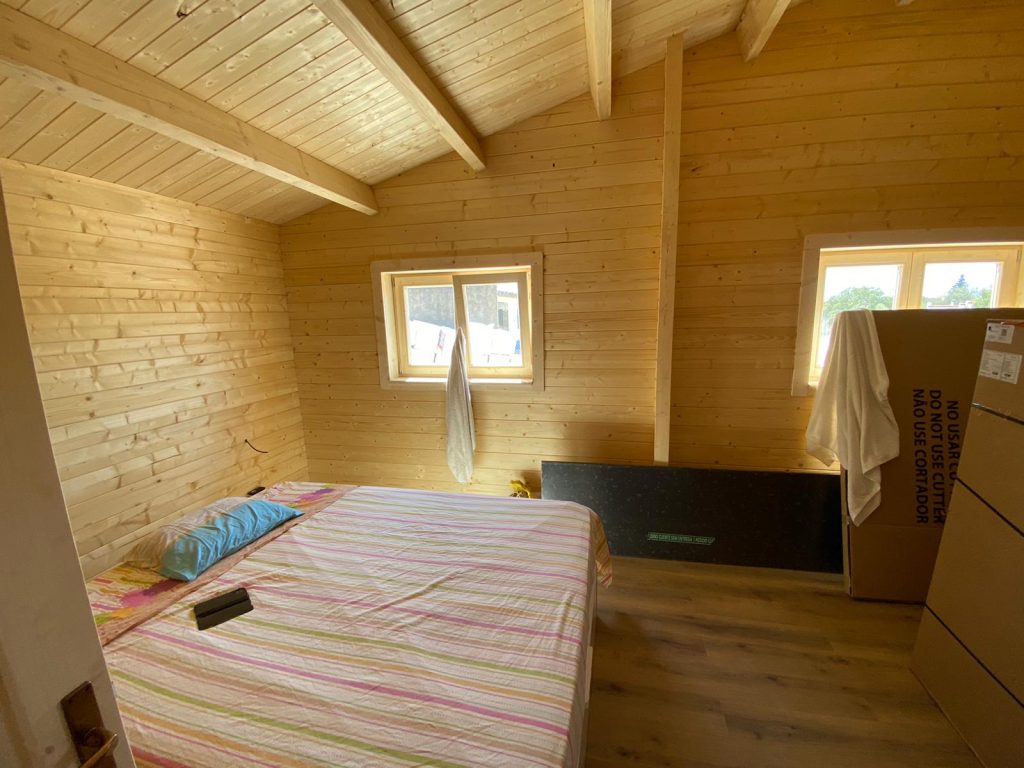 Quarto de casa de madeira com teto inclinado em pinho, vigas à vista e janelas em madeira