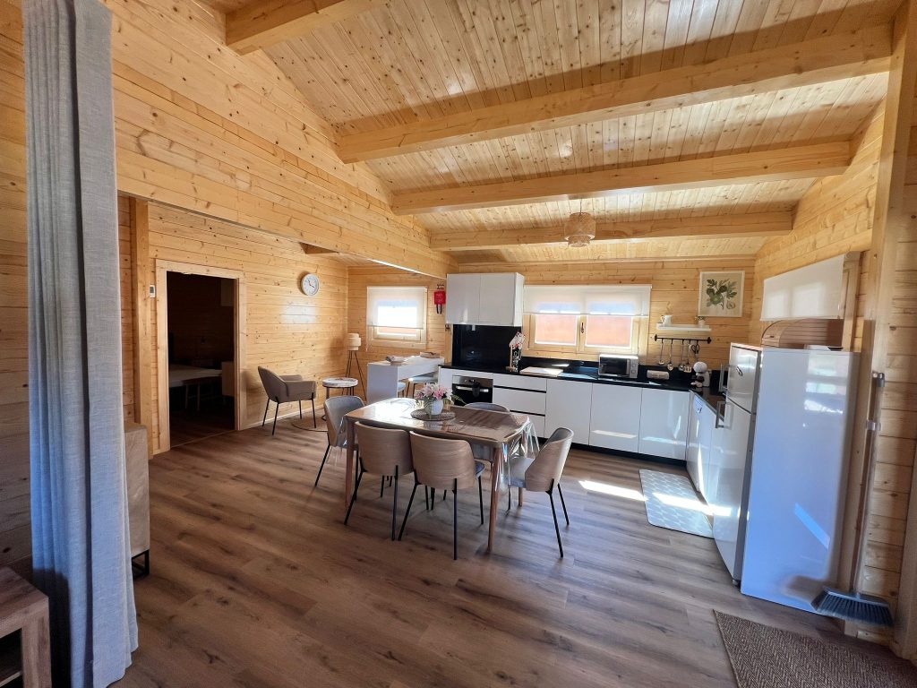 Open space com cozinha, ilha e mesa de jantar numa casa de madeira maciça com teto em pinho e vigas à vista